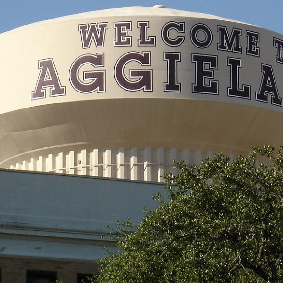 The Arbors at Wolf Pen Creek apartments Water tower with "WELCOME TO AGGIELAND" and Texas A&M logo, partially obscured by greenery.