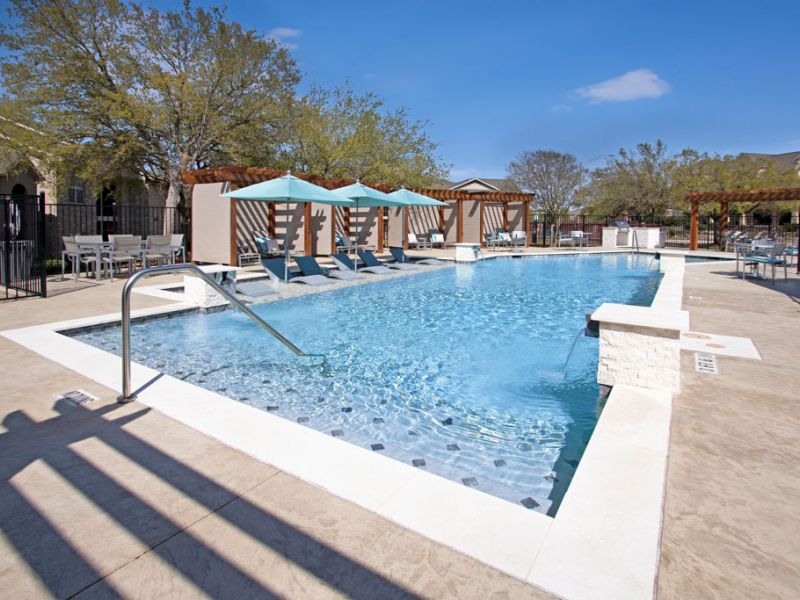 The Arbors at Wolf Pen Creek apartments Outdoor pool with lounge chairs, pergolas, and trees in the background under a clear blue sky.