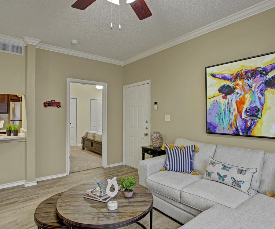 The Arbors at Wolf Pen Creek apartments Living room with a beige couch, colorful cow painting, ceiling fan, and view into the kitchen and bedroom.