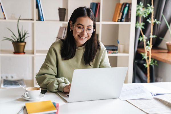 The Arbors at Wolf Pen Creek apartments resident in glasses smiling while working on a laptop at a desk with a cup of coffee and books in the background.