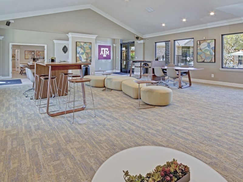 The Arbors at Wolf Pen Creek apartments Modern lounge area with seating, tables, and bar stools. Large windows and Texas A&M logo on the wall.