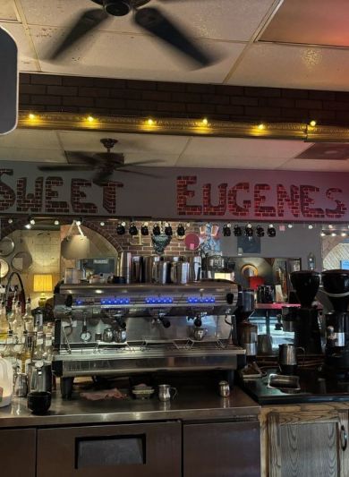 Coffee shop counter with espresso machines, syrup bottles, and a sign reading "SWEET EUGENES" in the mirror near The Arbors at Wolf Pen Creek apartments.