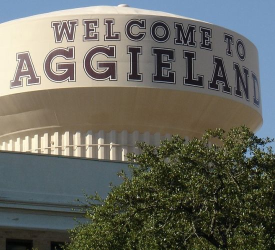 The Arbors at Wolf Pen Creek apartments Water tower with "WELCOME TO AGGIELAND" and Texas A&M logo, partially obscured by greenery.
