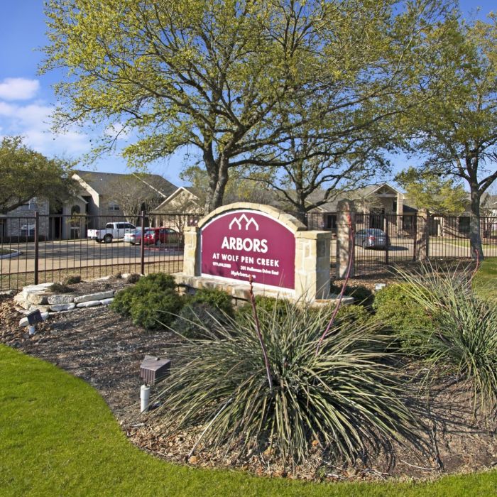 Entrance to Arbors at Wolf Pen Creek, featuring landscaped greenery and a maroon sign under a blue sky.