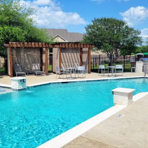The Arbors at Wolf Pen Creek apartments Outdoor pool with lounge chairs and pergolas, surrounded by trees and clear blue sky.